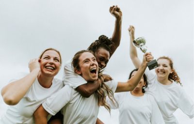 An excited team of women soccer athletes laugh and hug each other holding a championship trophy.